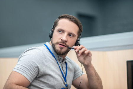 Work moment. Engaged serious man in gray tshirt and badge wearing headphones listening and speaking...