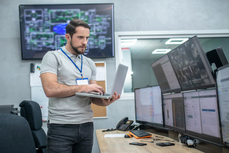 Attention and focus. Attentive young muscular man with badge and laptop standing in front of computer monitorsの写真素材