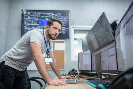 Great responsibility. Serious young adult man with badge leaning over table watching multiple monitors at same timeの写真素材