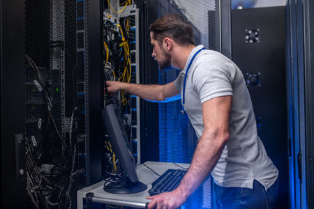 Server checking. Young bearded man in tshirt and jeans with badge in server room near open cabinet...