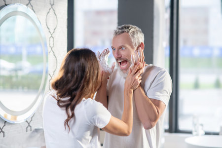 Cheerful morning. Grimacing cheerful bearded husband with raised hands in soapy foam and wife touching himの写真素材