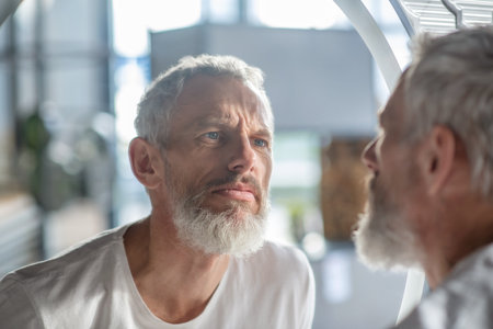 Looking at the reflection. A bearded grey-haired man looking at the mirror