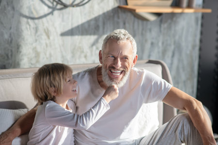 Fun game. Cheerful child and gray-haired bearded man sitting on bed playing laughing on sunny day