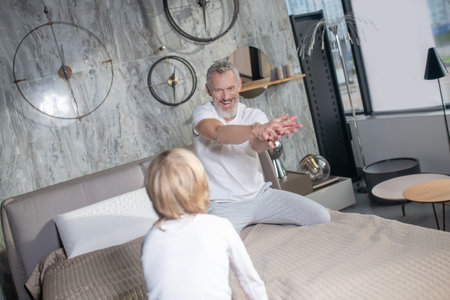 Interesting entertainment. Gray-haired bearded joyfully smiling man outstretched arms and child opposite sitting on bedの写真素材