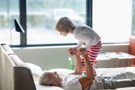 Time with dad. Adult strong dad lying on bed in pajamas raising child leaning on his armsの写真素材
