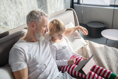 Viewing, interesting. Attentive bearded man and child in pajamas looking together intently at tablet...