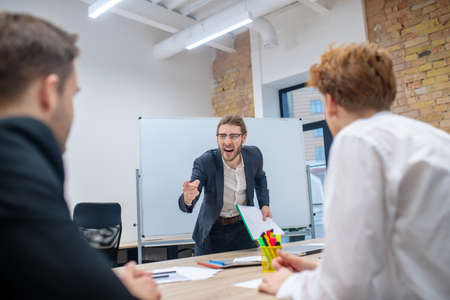 Angry manager. Angry man with glasses in a dark suit standing shouting at employees sitting at table
