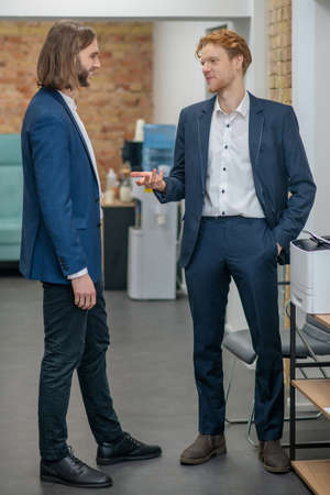 Communication, employees. Two smiling young adult men in dark suits communicating in office space in...