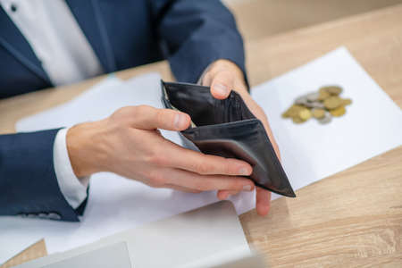 Lack of money. Hands of man in business suit with open wallet over table next to small coins,...