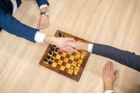 Handshake, players. Hands of two men in suits shaking hands over chessboard with figures in front of...