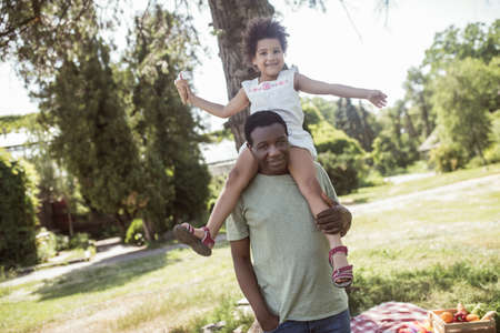 Happy family. Dark-skinned cute kid sitting on her dads shoulders