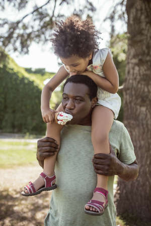 Happy family. Dark-skinned cute kid sitting on her dads shouldersの写真素材