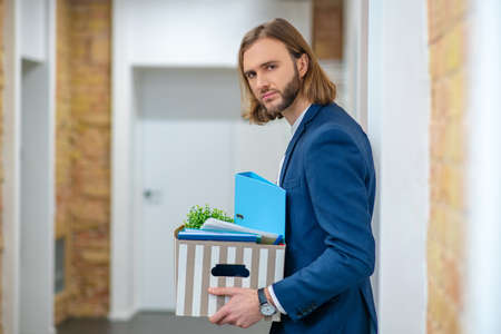 Working day, nuisance. Serious young long-haired man in blue suit standing with box in office corridorの写真素材