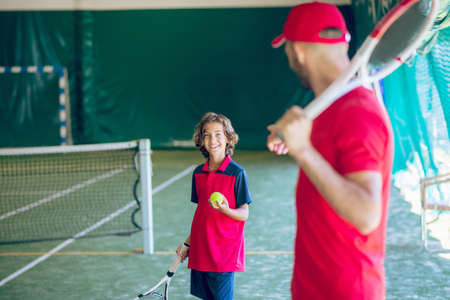 In gym. Young bearded man in a red cap holding a tennis racket and talking to a boyの写真素材