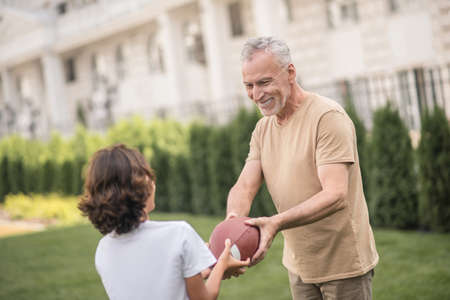 Time to play. Gray-haired dad giving a ball to his son and smiling