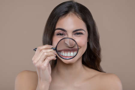 Healthy teeth. Young cheerful woman with bared shoulders holding magnifying glass near smiling mouth...