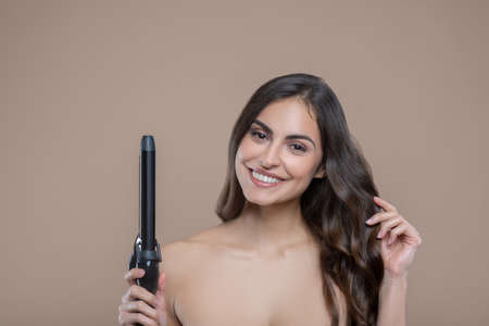 Curling iron. Half-naked beautiful young woman with curling iron in one hand with other touching her...