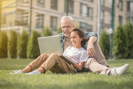 Connected. Son and dad sitting together on the grass with a laptop and looking contented