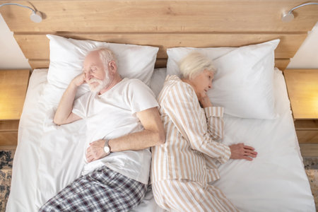 Trying to sleep. Aged married couple lying in bed back to backの写真素材