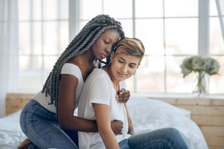 In love. Dark-skinned girl hugging her short-haired girlfriend from the back and looking tenderの写真素材