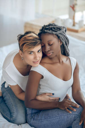 Closeness. Short-haired girl hugging her dark-skinned girlfriend from the back and looking pleasedの写真素材