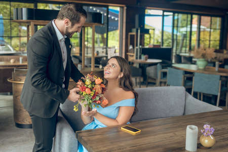 Nice meeting. Smiling caring loving man in suit with flowers and happy woman sitting in cafeの写真素材