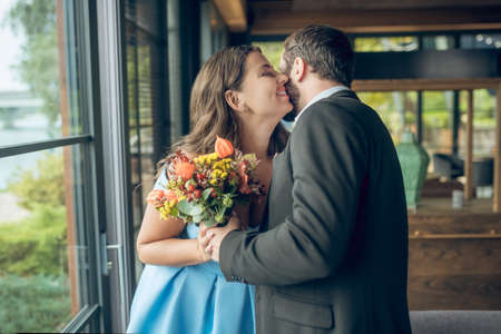 Happy couple. Man in suit with flowers and happy pretty long-haired woman in light blue dress are standing hugging in cafeの写真素材