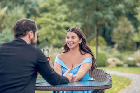 Happy together. Shining young pretty woman with glass of wine and man touching hand sitting chatting in outdoor cafeの写真素材