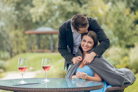 Love, care. Young adult man in dark suit with plaid carefully covering happy pretty woman sitting in cafe on natureの写真素材