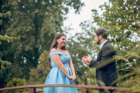 Engagement. Attentive man in suit with red small box and joyful shining woman in dress standing...