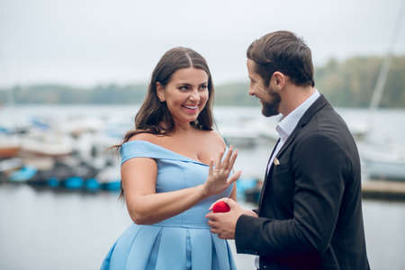 Significant event. Joyful man with small red box proposing and agreeing beautiful woman showing ring on her fingerの写真素材