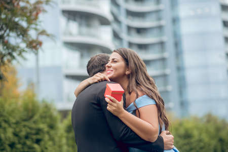 Lucky day. Happy young long-haired woman receiving gift hugging man in dark suit standing outdoors on summer dayの写真素材