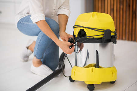 Yellow vacuum cleaner. Woman in jeans and white loafers crouching near yellow vacuum cleaner holding black cord without faceの写真素材