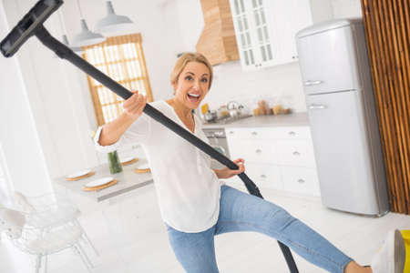 Happy moment. Energetic cheerful adult woman with raised brush of vacuum cleaner and leg in kitchenの写真素材
