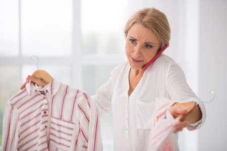 Indecision. Indecisive adult long-haired woman with clothes in hands talking on smartphone in bright...