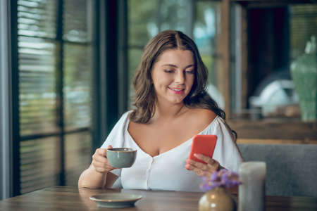 Interesting message. Young joyful smiling woman with cup of coffee looking interested in smartphone...