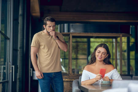 Unexpected moment. Smiling woman sitting at table looking at smartphone and surprised man with hand near face standing from behindの写真素材