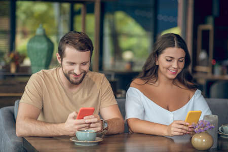 Interesting information. Young adult smiling man and woman in casual clothes looking at their smartphones at table in cafeの写真素材