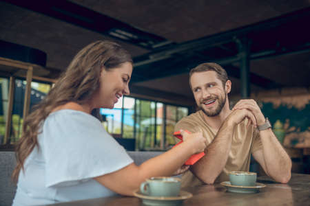 Joyful moment. Young shining woman in white blouse opening gift and joyful bearded man in love sitting in summer cafeの写真素材