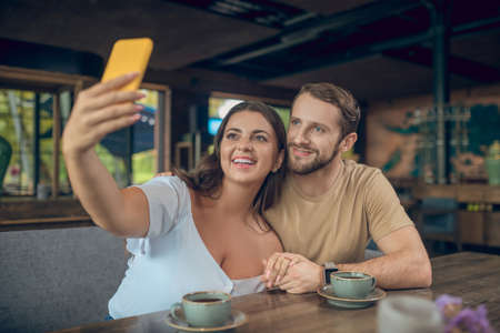 Successful selfie. Smiling lovers woman and man taking selfie on smartphone while sitting in cafe in afternoonの写真素材