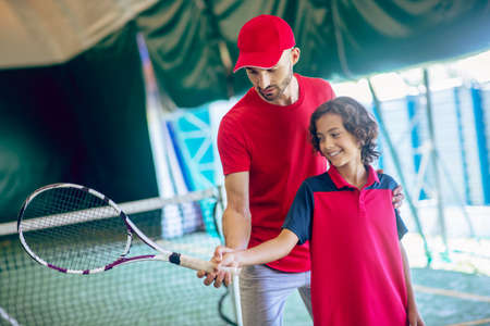 Learning to play tennis. Male tennis coach in red clothes showing a boy how to hold the racket