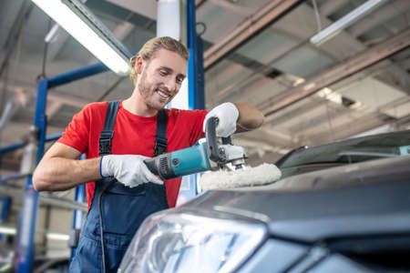 Auto repair shop. Young bearded specialist man in red tshirt and overalls improving appearance of...