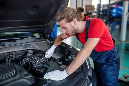 Car repair. Young adult man in uniform with wrench leaning over open hood of car in garage