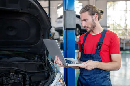 Responsibility, work. Serious attentive young adult man standing with computer near car in garageの写真素材