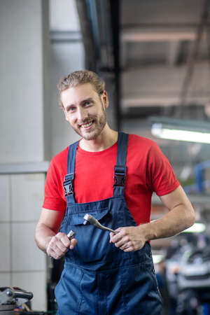 Workday, repairman. Happy smiling young male repairman in blue overalls and red tshirt with iron detail in handsの写真素材