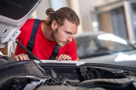 Responsibility, attentiveness. Attentive young bearded repairman bending low and attentively examining open hood in workshopの写真素材