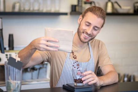 Favorite work. Joyful young bearded man in apron having fun pouring coffee beans into measuring glass behind cafe counterの写真素材