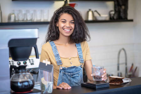 Working day, cafe. Happy young mulatto woman with coffee beans in glass standing behind counter in coffee shopの写真素材