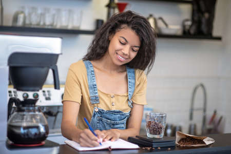 Accounting, cafe. Pretty young mulatto woman writing weight of coffee in notebook at bar counter in...