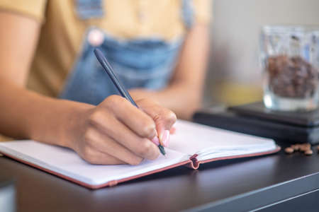 Accounting. Womans thin graceful hand writing with pen in notebook weight of coffee beans on counter